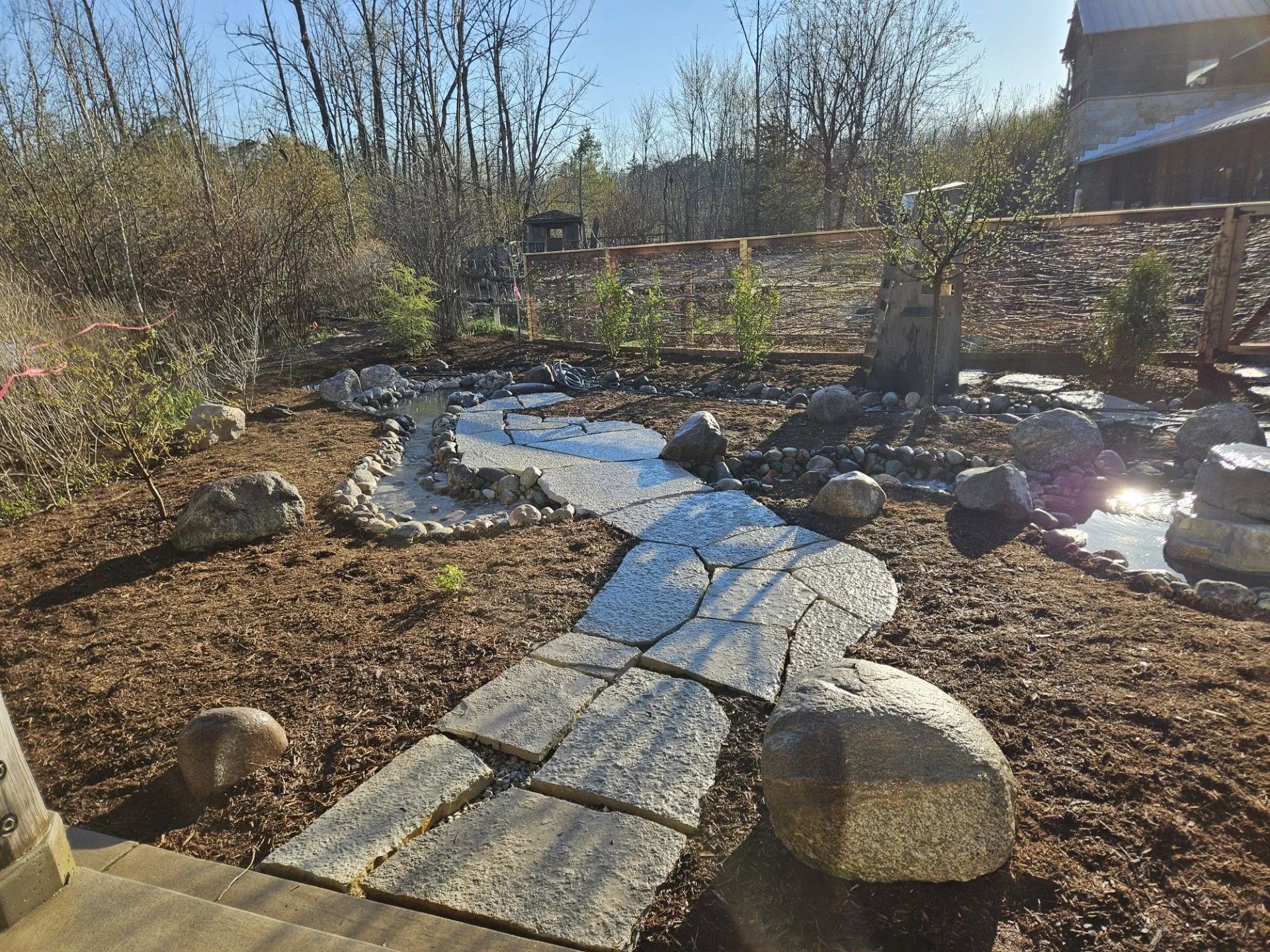 A stone walkway leads through a mulched garden area toward a pond with surrounding rocks and a wooden fence background.