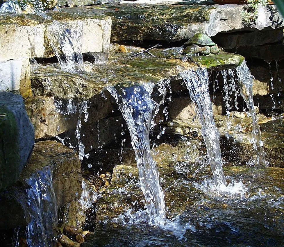 A small, stone-tiered garden waterfall with a decorative turtle statue sitting on the ledge.