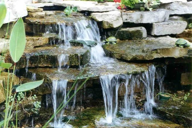 A tiered stone waterfall cascading down layers of flat rocks into a pond, featuring two small turtle figurines.