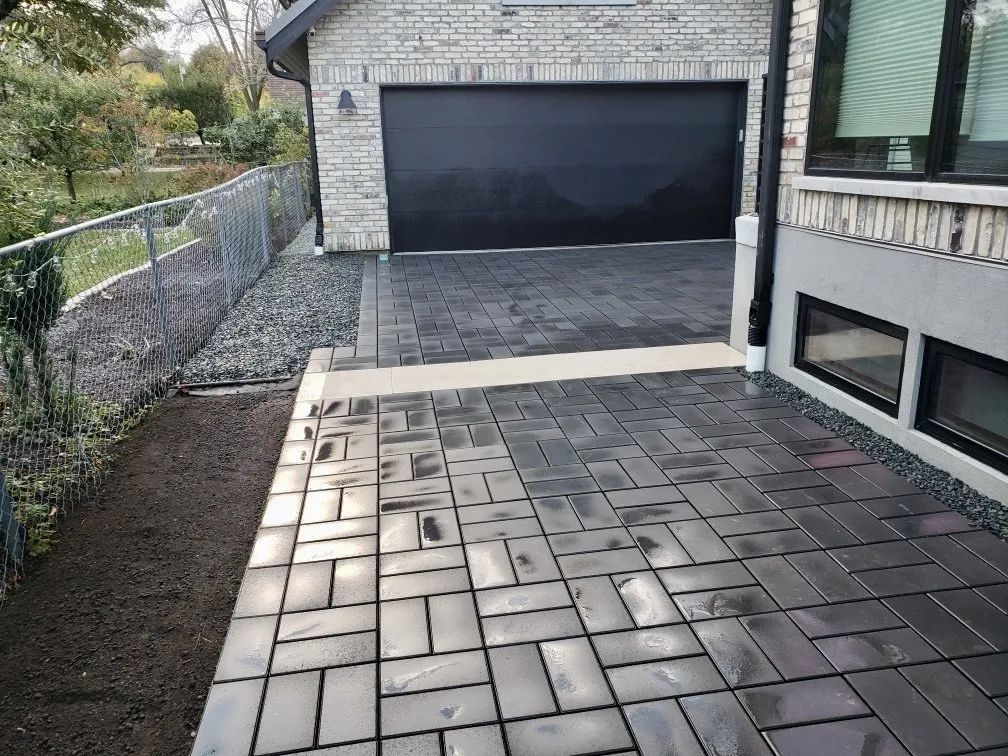 A grey stone patio made of rectangular pavers leading to a dark garage door next to a light-colored brick house.