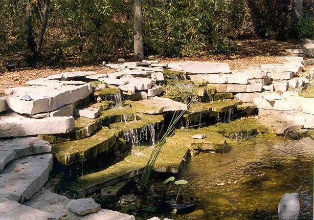 A multi-tiered stone waterfall cascades into a small garden pond surrounded by trees and leaf litter.