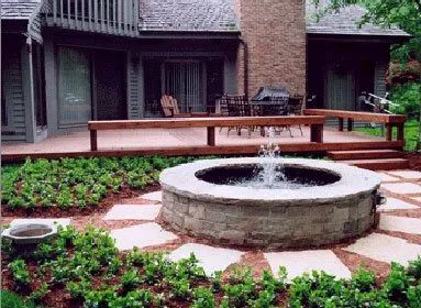 A circular stone fountain sits in a garden bed in front of a house with a wooden deck and stone chimney.