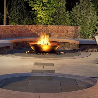 A modern metal fire bowl glows with fire at night in a stone-paved patio area with built-in curved stone seating.
