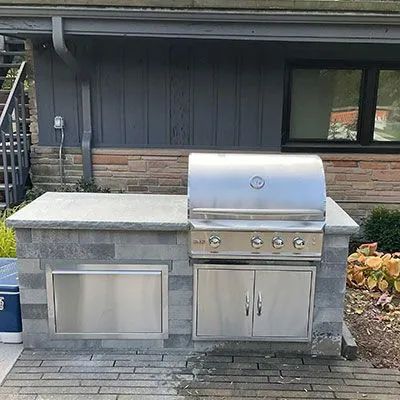 Outdoor kitchen island with a stainless steel built-in grill and storage doors against a stone-walled house exterior.