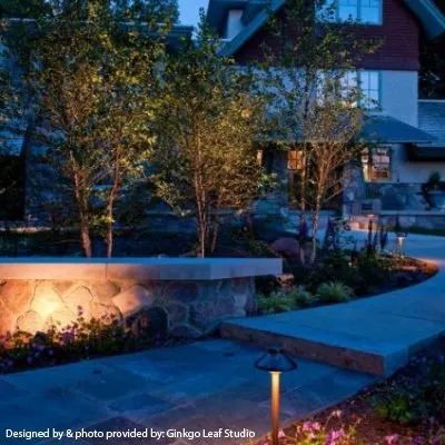 A stone garden wall and paved path illuminated by warm outdoor landscape lighting in front of a house at dusk.