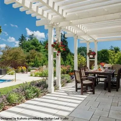 A white wooden pergola shades a stone patio with outdoor dining furniture, lush greenery, and a nearby swimming pool.