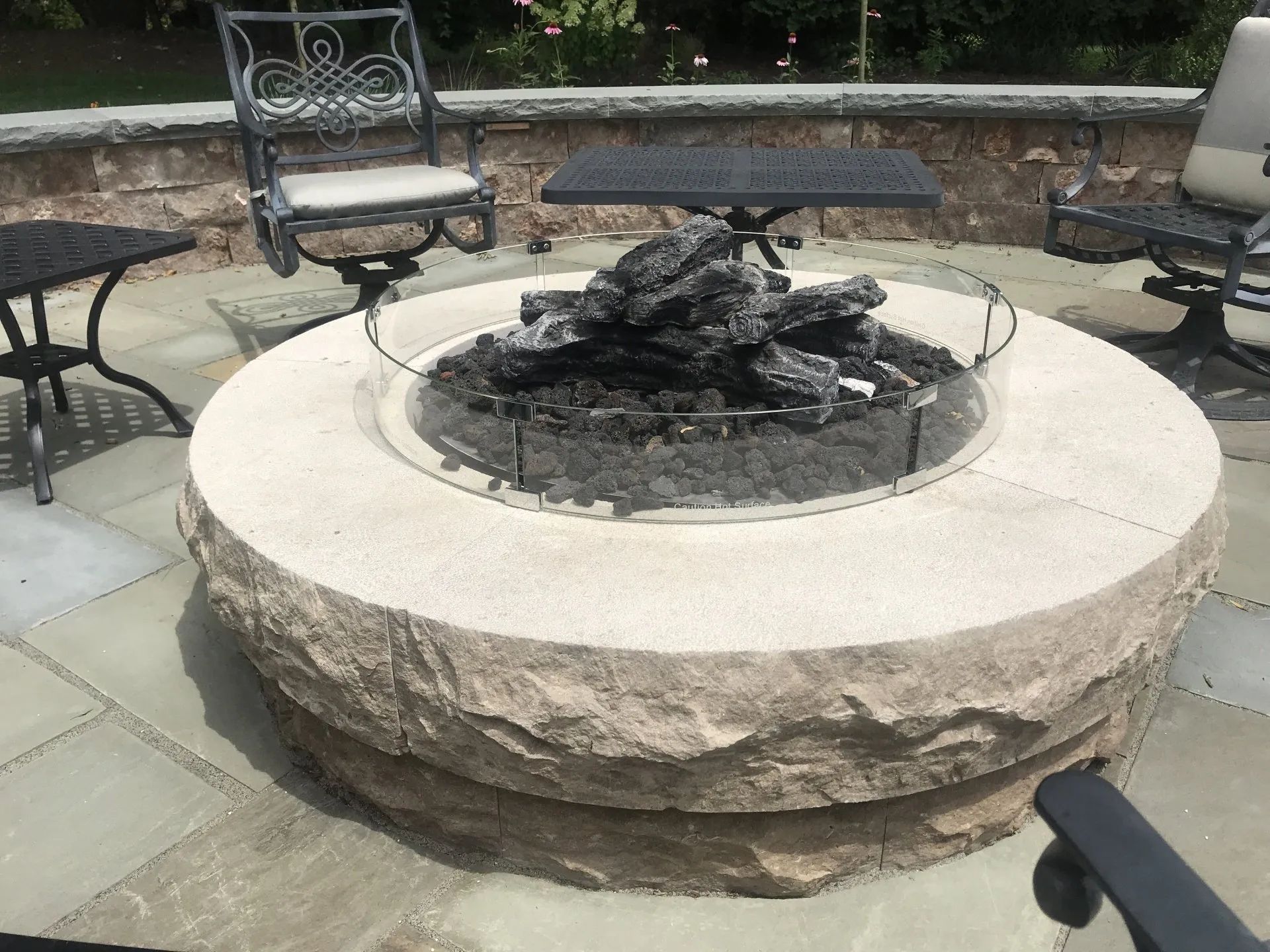 A circular outdoor fire pit with a natural stone base and glass wind guard, situated on a stone patio with chairs.