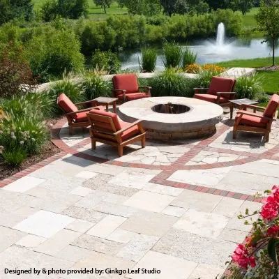 A stone patio with a central circular fire pit and four lounge chairs, overlooking a pond with a fountain.