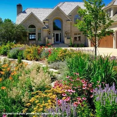 A two-story stone house with a shingled roof, set behind a lush garden filled with colorful wildflowers and green shrubs.