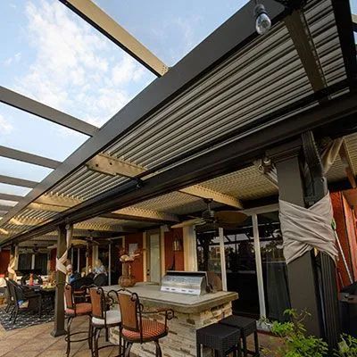 A covered outdoor patio featuring a stone bar counter, stools, and a pergola with adjustable slats on a sunny day.