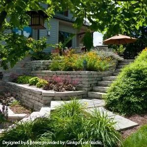 A terraced stone staircase leads up to a house with lush flower beds and greenery, framed by trees and a patio umbrella.