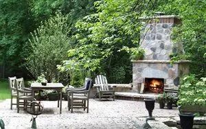 A stone outdoor fireplace with a lit fire stands on a paved patio beside a wooden dining table and chairs in a wooded area.