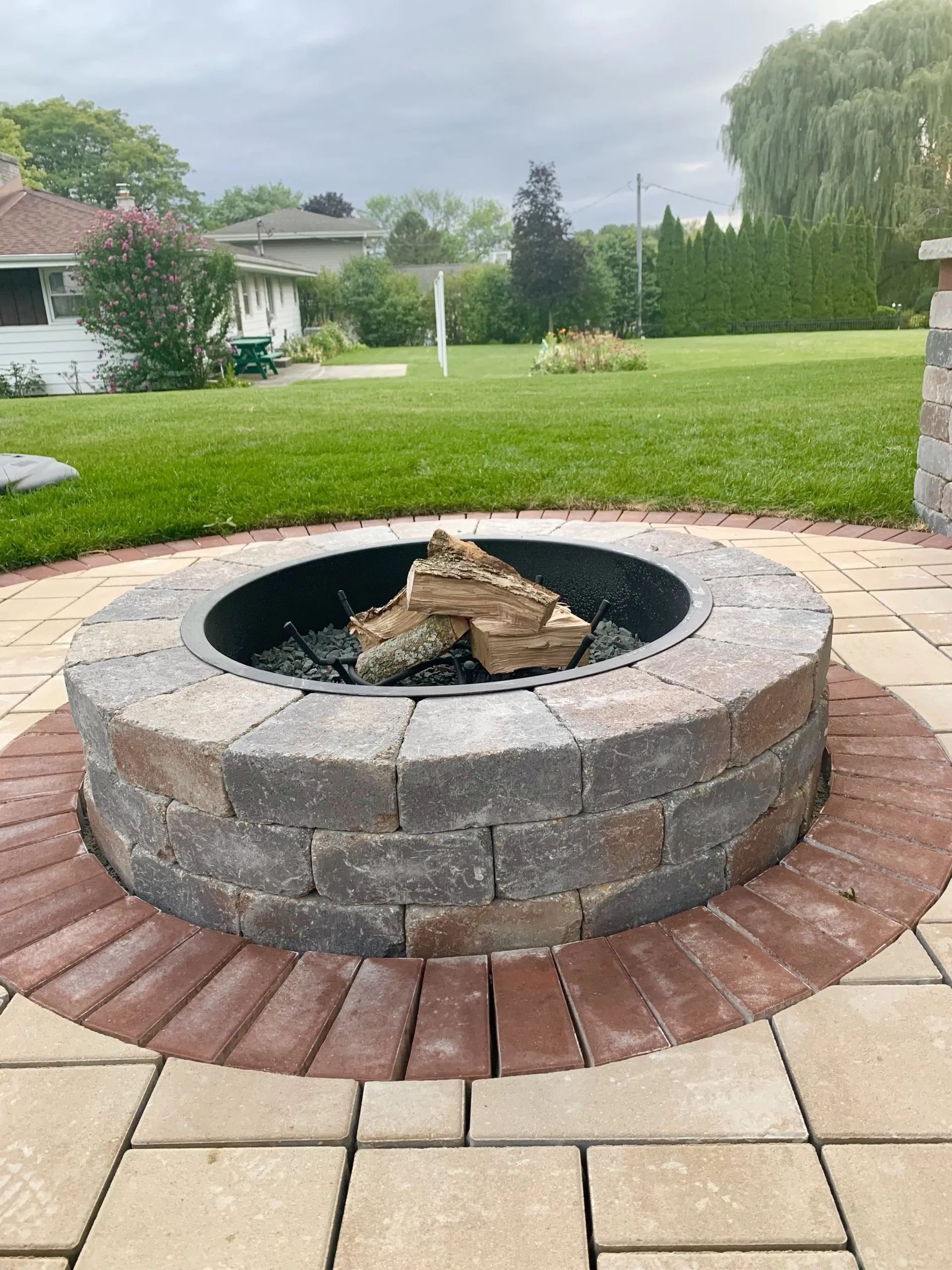 A circular stone fire pit sits on a paved patio in a grassy backyard with a house and trees in the distance.