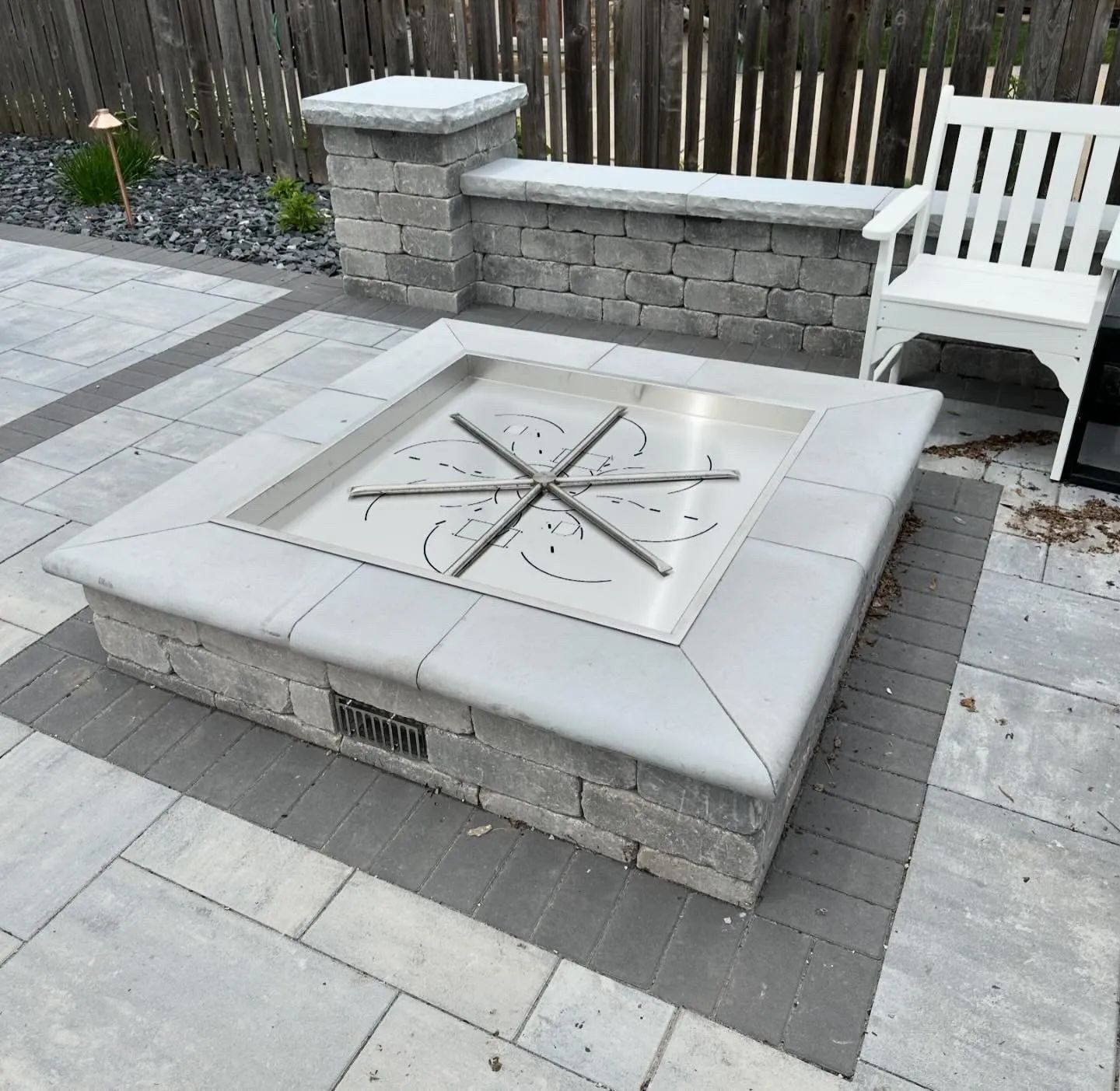 A square stone fire pit with a star-shaped gas burner set into a paved patio near a low stone wall and white chair.