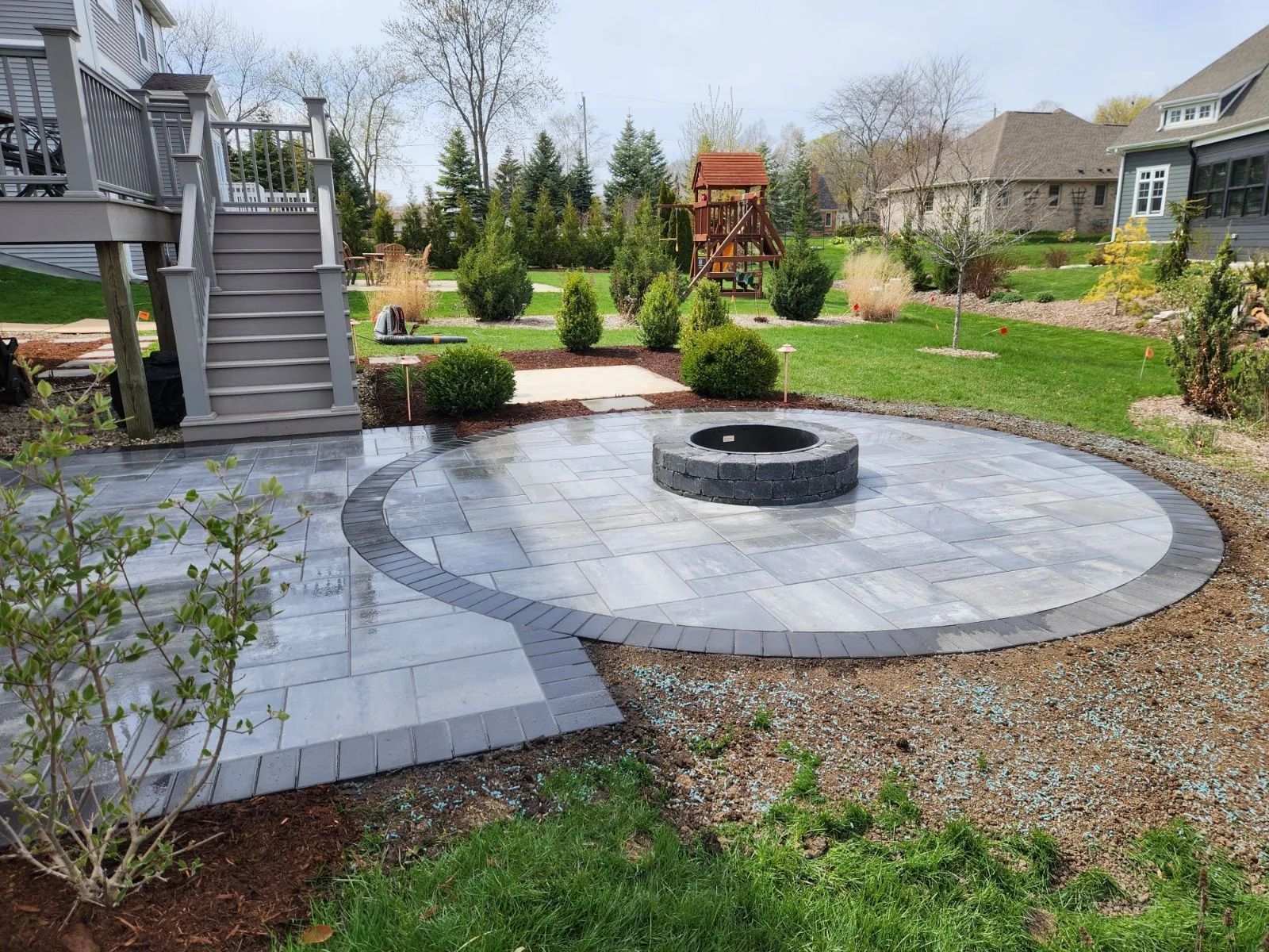 A backyard patio with a circular fire pit, stone pavers, and stairs leading to a raised wooden deck on a sunny day.