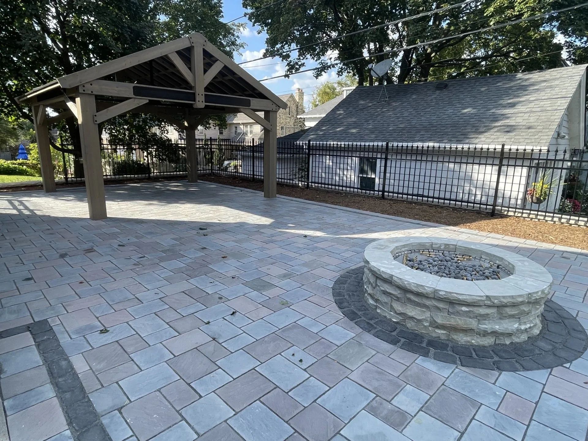 A backyard patio with light-colored pavers, a stone fire pit, and a wooden pavilion structure under trees.