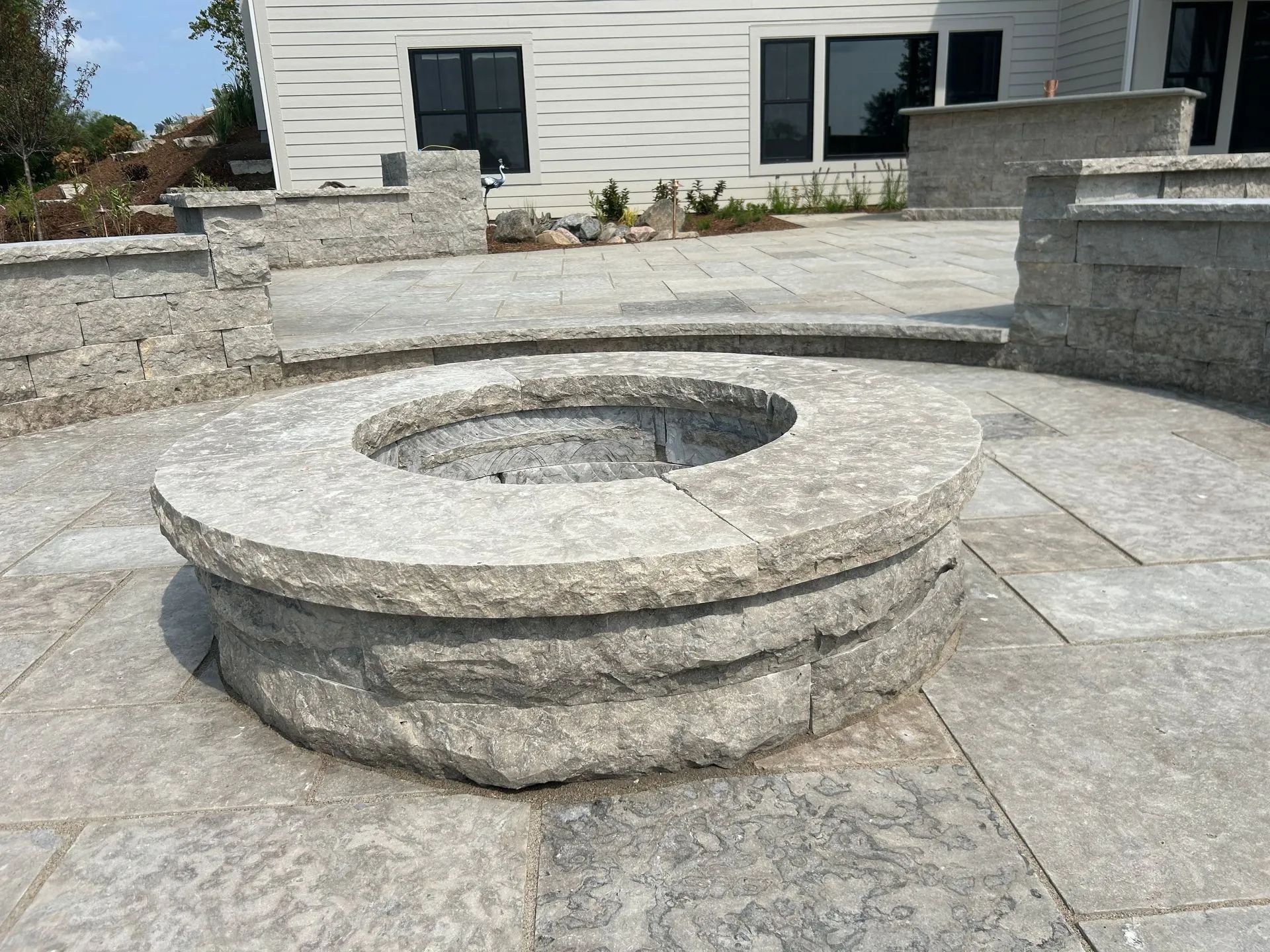 A circular, light gray stone fire pit sits on a matching stone patio in front of a modern house.
