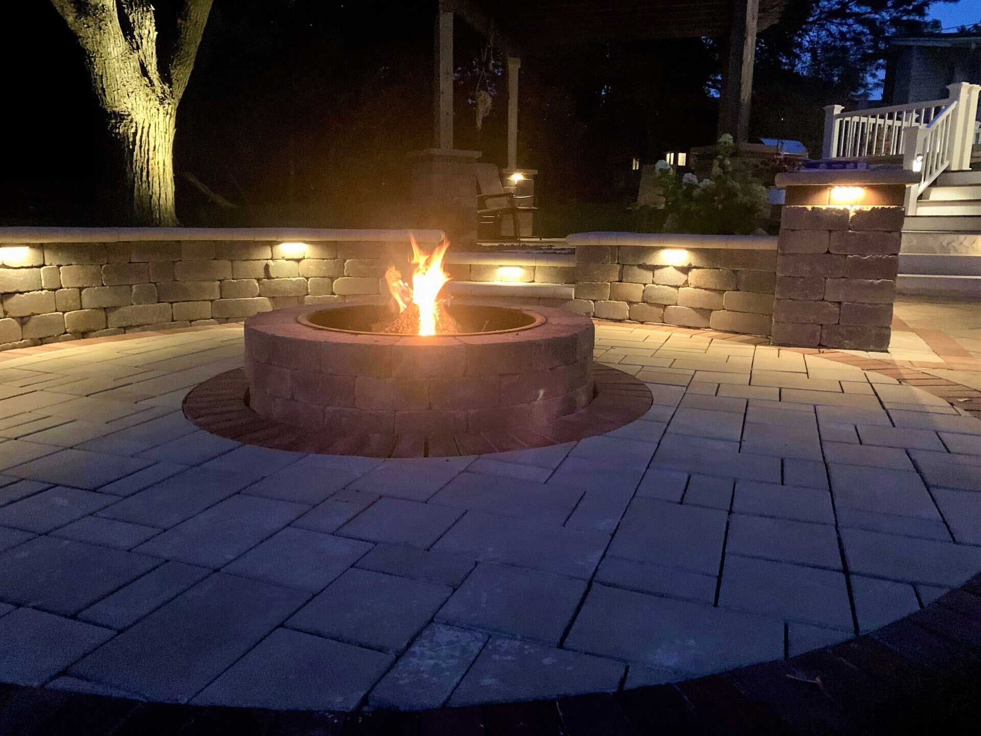 A circular stone fire pit with a glowing flame in the center of a paved stone patio at night, illuminated by accent lights.
