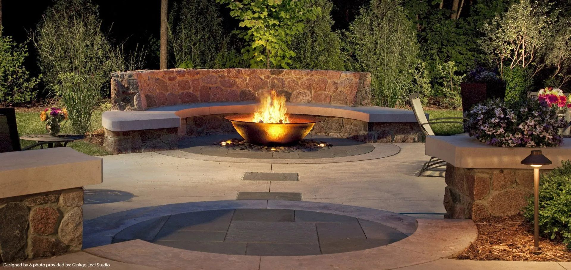 A backyard stone patio at night, featuring a circular fire bowl, curved built-in seating, and landscape lighting.