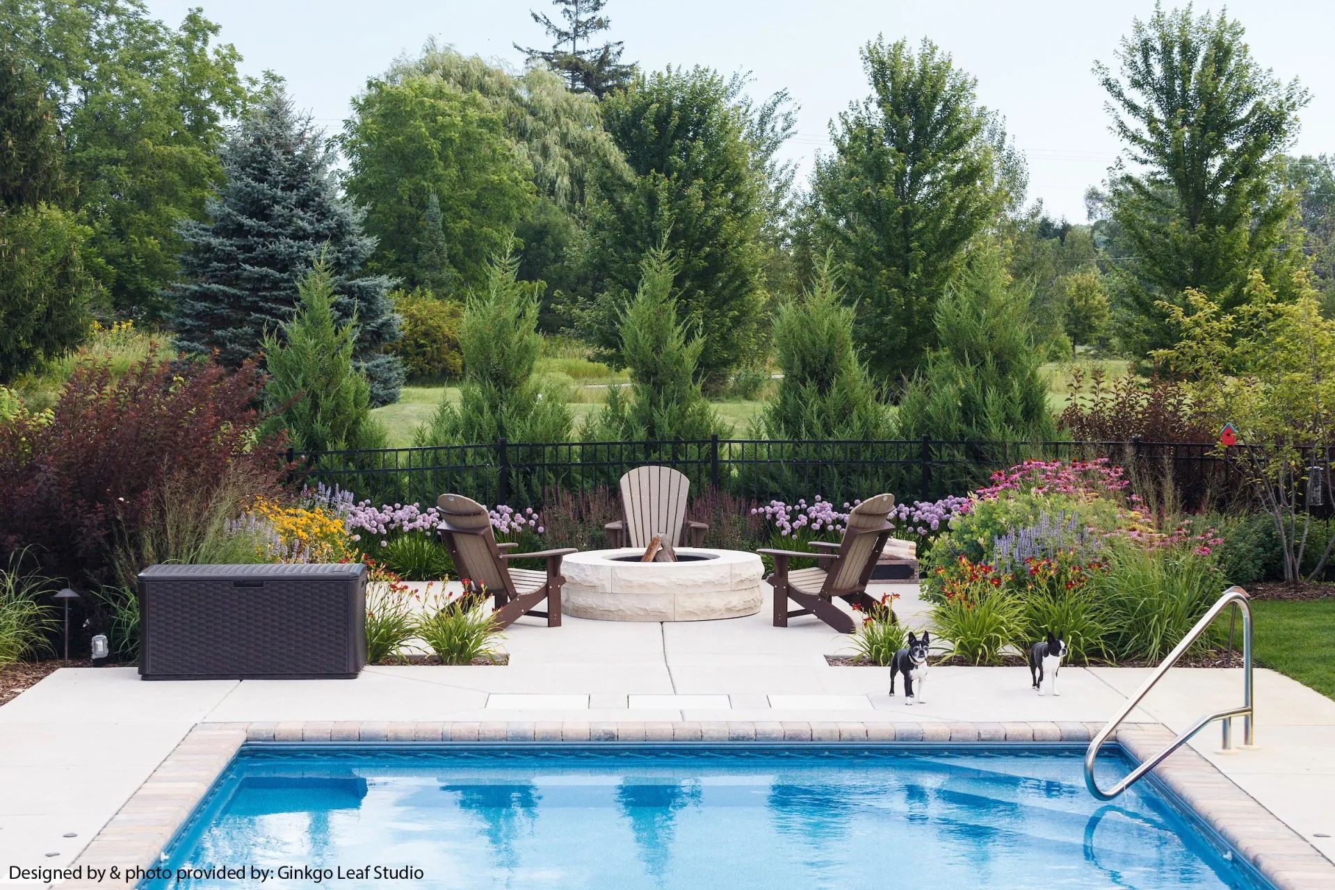 A swimming pool in a lush backyard patio featuring a fire pit with Adirondack chairs, trees, and landscaping.
