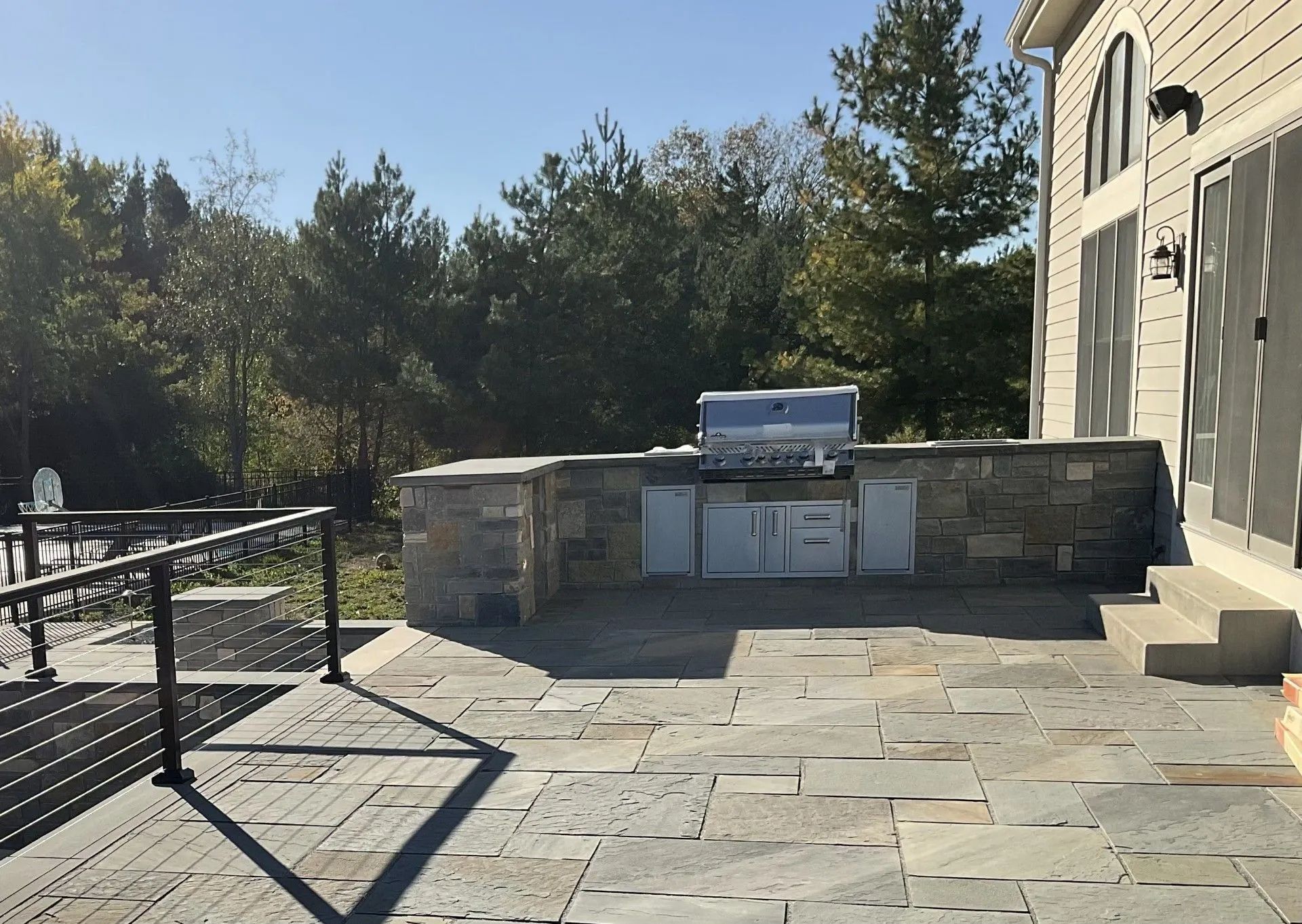 An outdoor stone patio features a built-in stainless steel grill station, a metal railing, and steps leading to a house.