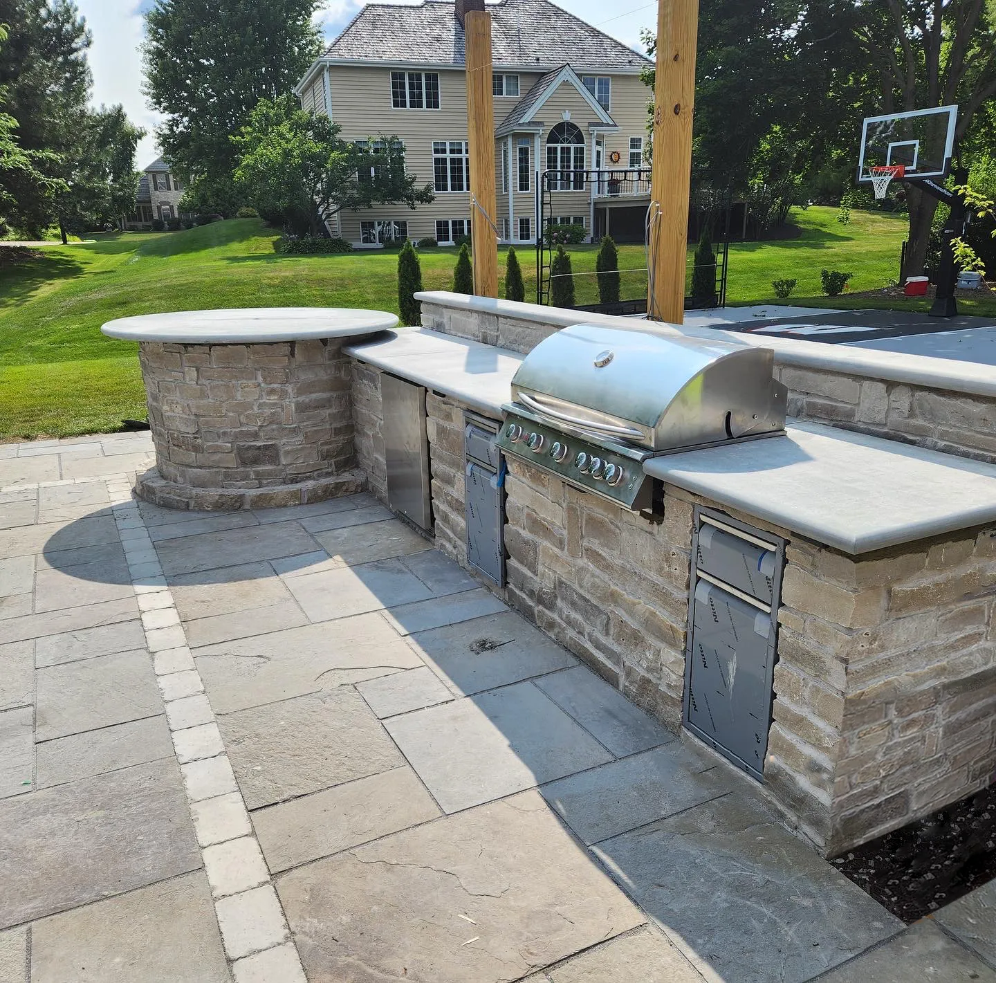 A stone outdoor kitchen with a built-in grill and circular counter on a patio, with a house in the background.