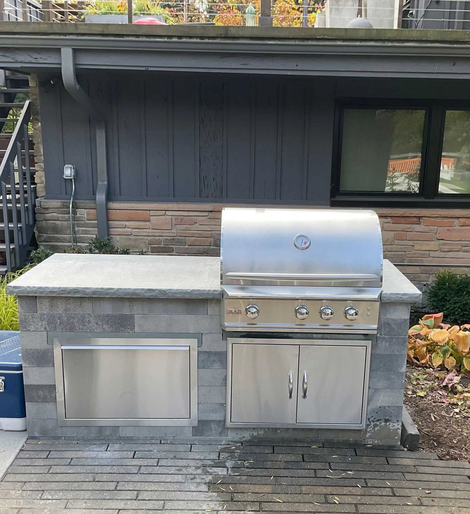 An outdoor kitchen island with a stainless steel grill, cabinets, and a stone countertop on a patio.
