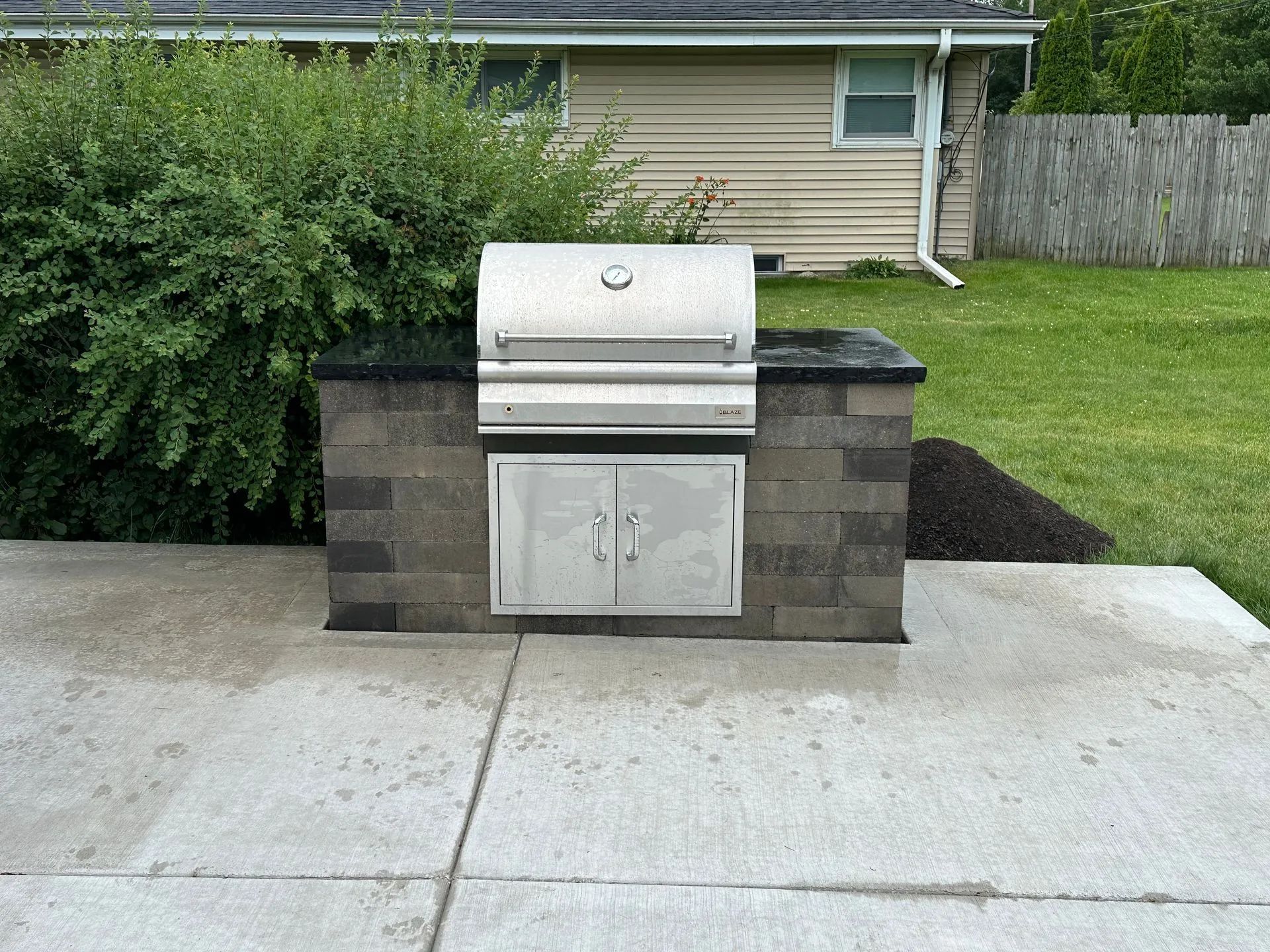 A stainless steel built-in grill set into a stone block counter on a concrete patio in a grassy backyard.