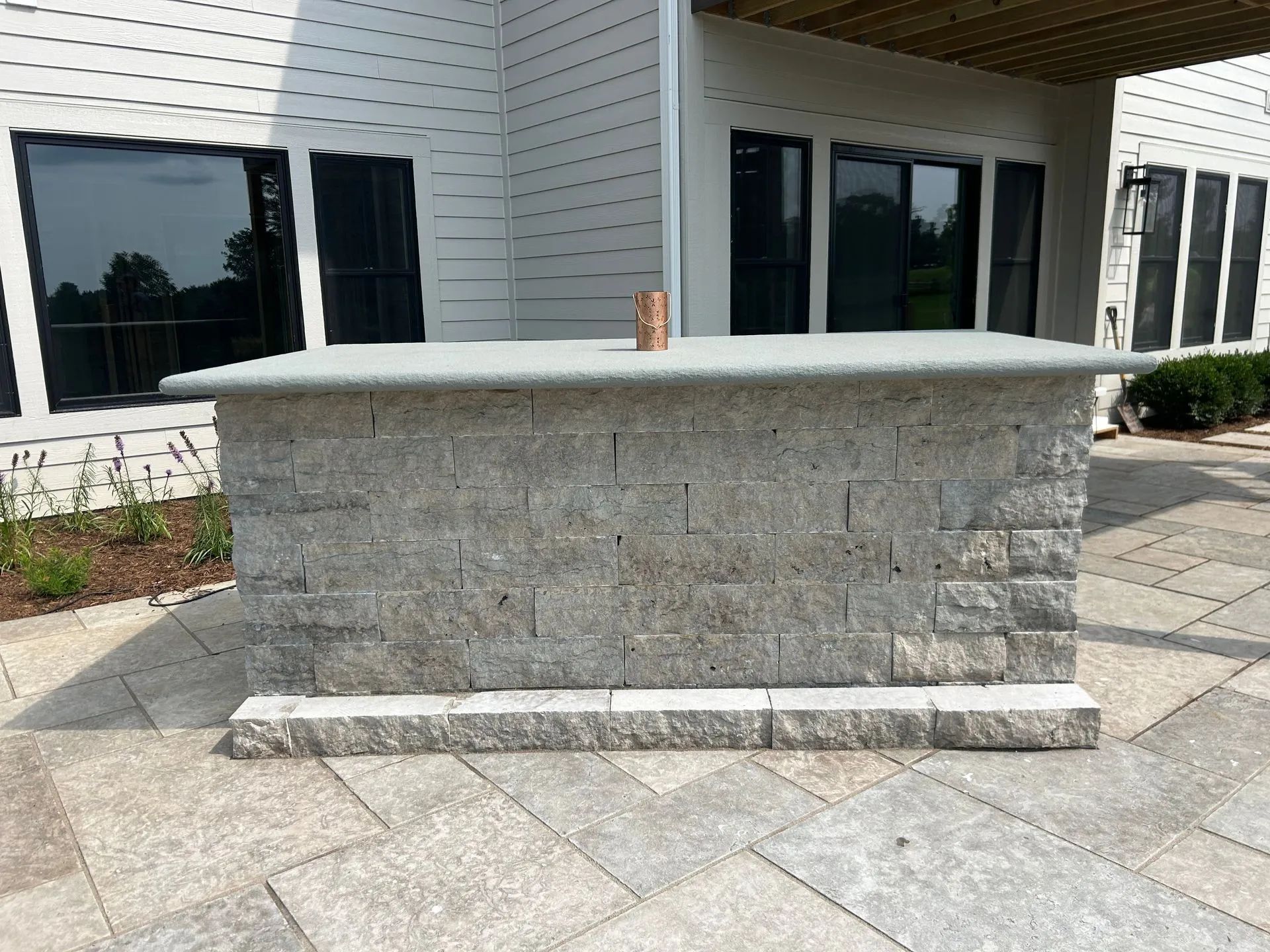 A rectangular, gray stone outdoor bar sits on a stone patio in front of a white house with large windows.