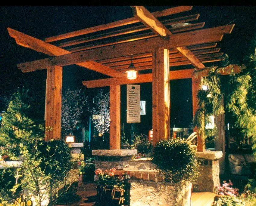 A wooden pergola with a hanging lantern stands over stone planters and garden greenery at night.