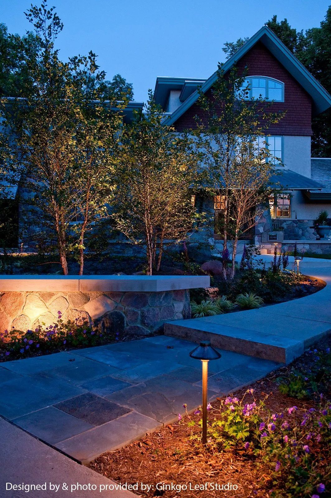 A stone-paved walkway at dusk, illuminated by small path lights and glowing spotlights directed at nearby trees.