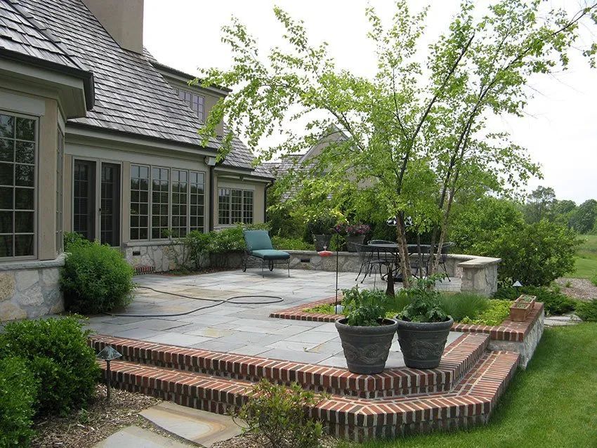 A stone patio featuring brick steps, potted plants, and a small tree, set against the side of a large suburban home.