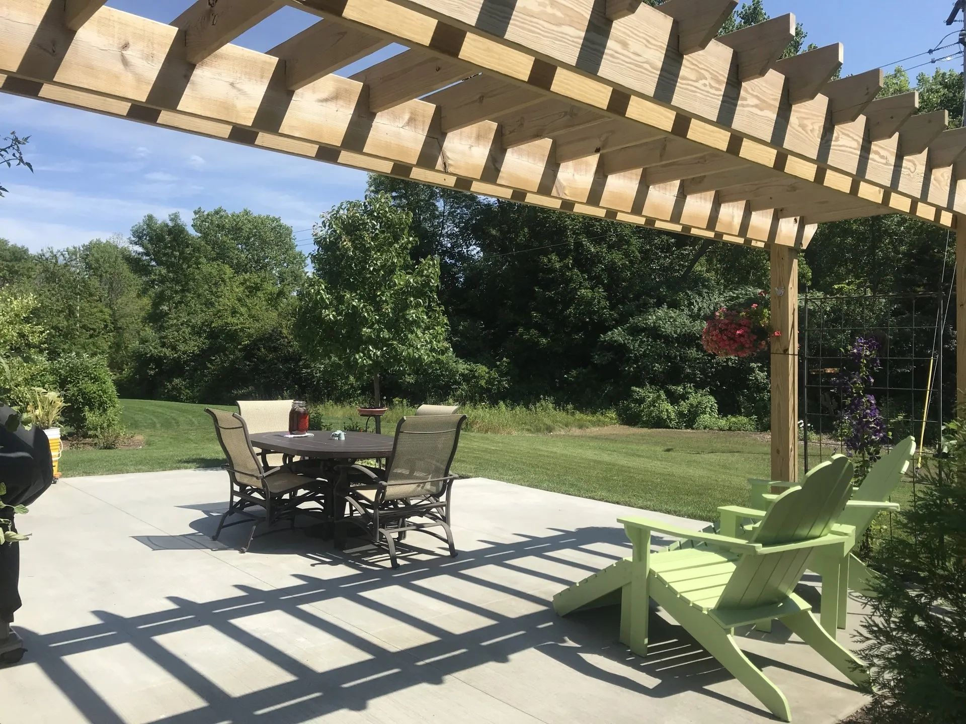 A concrete patio with a wooden pergola, a dining table set, and two lime green Adirondack chairs on a sunny day.