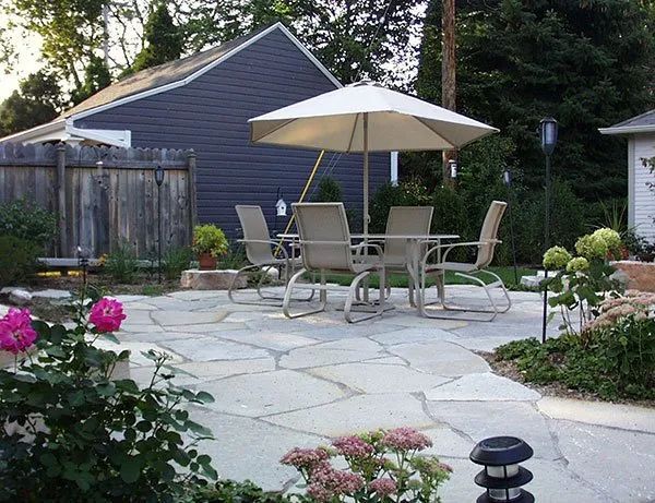 A beige patio umbrella over a dining set on a stone patio in a backyard with a blue shed and surrounding garden beds.