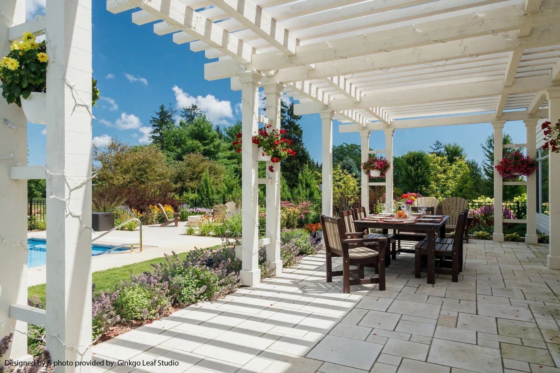 A white pergola shades a patio dining set next to a swimming pool, surrounded by lush landscaping and sunny blue skies.