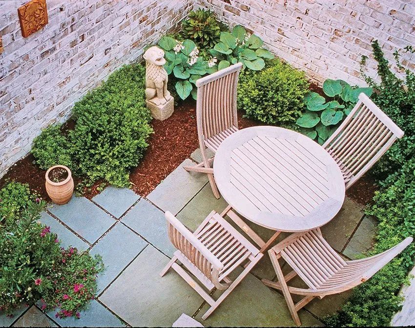 A small, square brick-walled courtyard featuring a round teak dining table, four folding chairs, and surrounding plants.