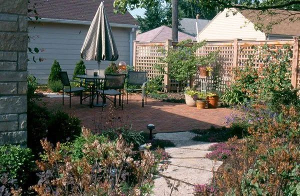 A brick patio with a small table, chairs, and a closed umbrella, surrounded by garden plants and a wooden lattice fence.