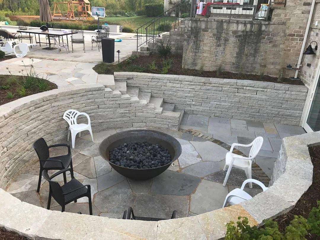 An outdoor stone fire pit area with a circular wall, flagstone patio, and several chairs arranged for seating.