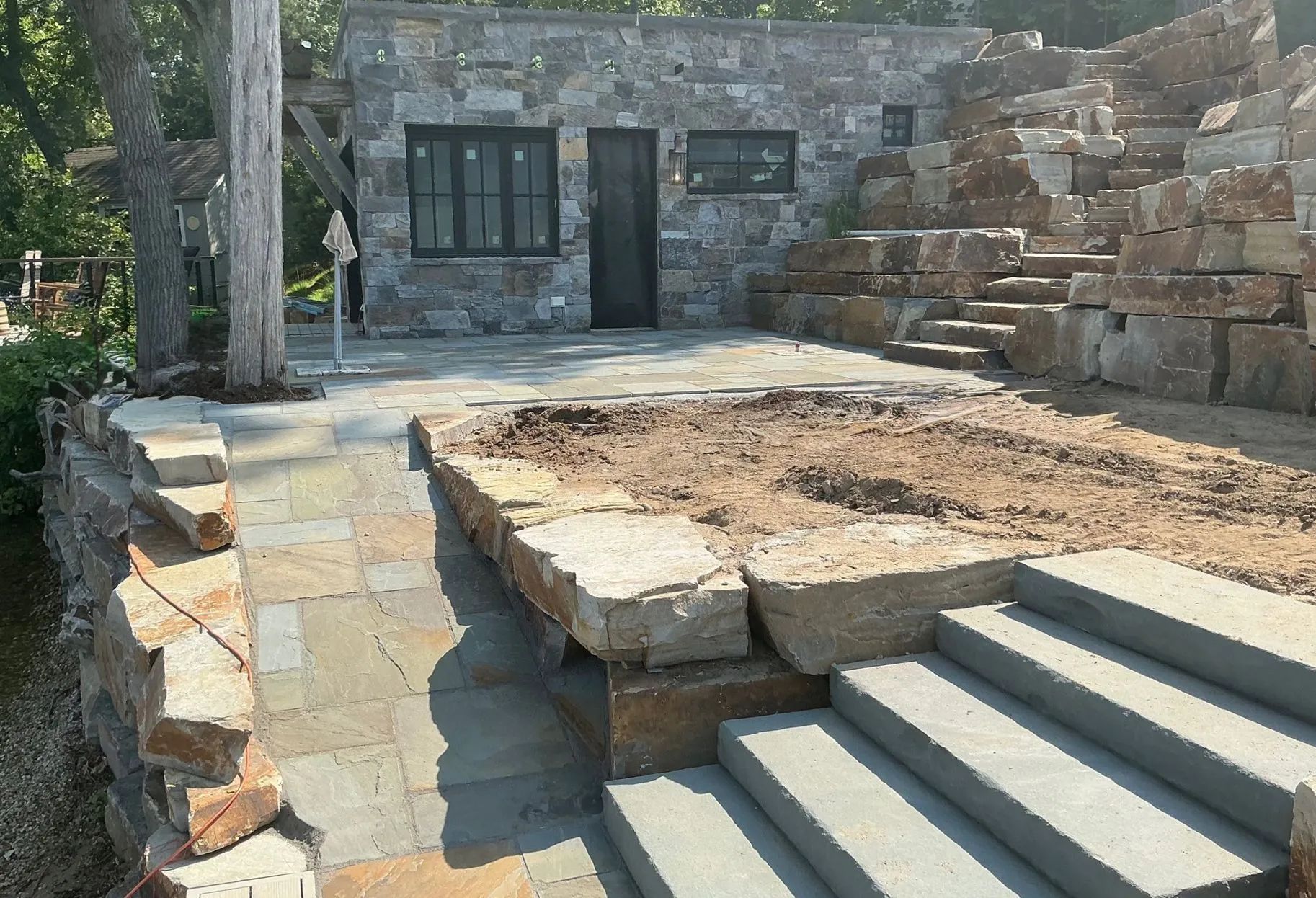 A stone building with a patio, stone retaining walls, and concrete stairs leading up to an unfinished landscape area.