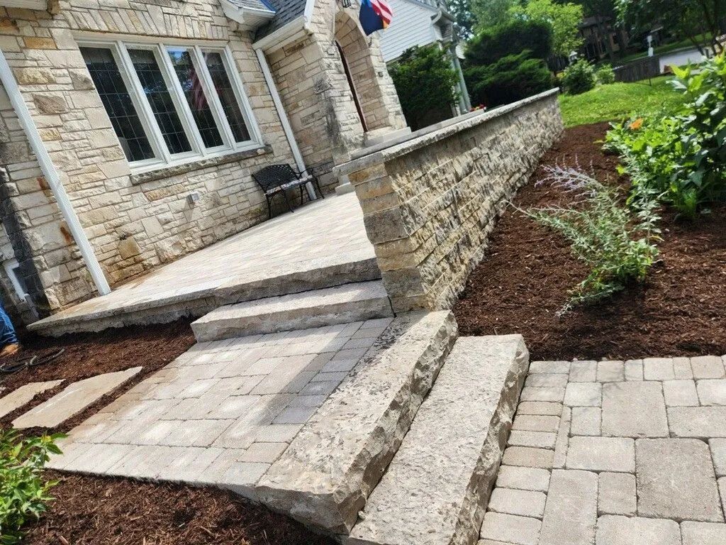 Stone steps leading to a light-colored stone house patio with a matching retaining wall and mulched garden beds.