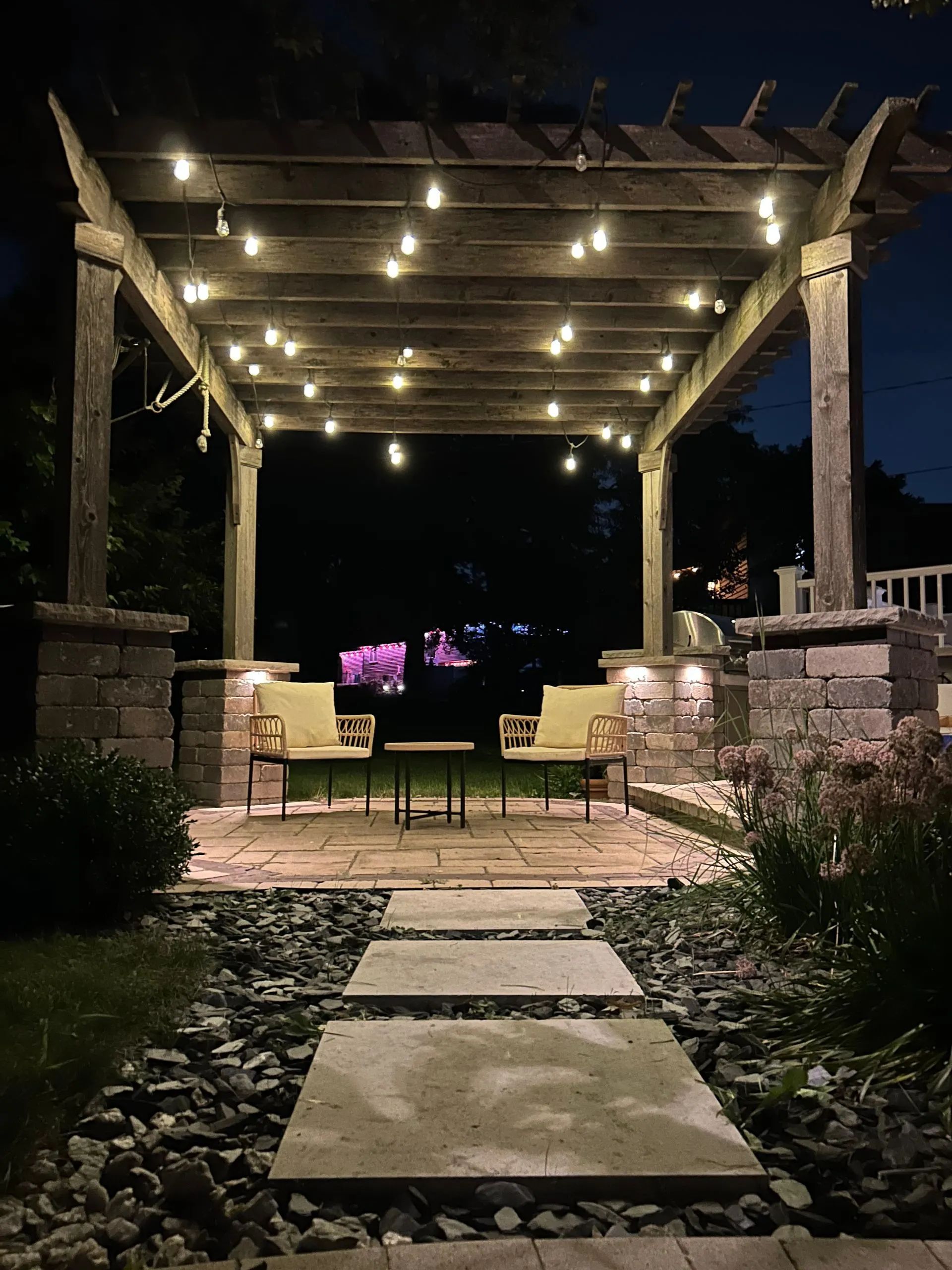 A cozy patio at night featuring a wooden pergola with string lights, two chairs, and a stone path leading to the area.