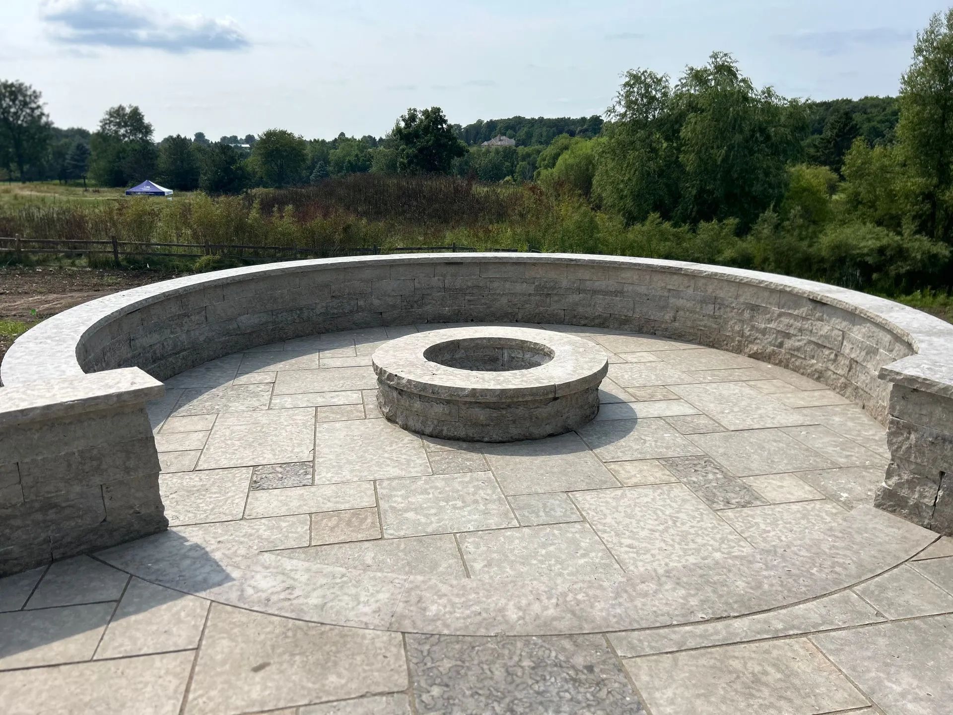 A circular stone fire pit sits on a matching stone patio enclosed by a curved stone bench in a grassy park setting.