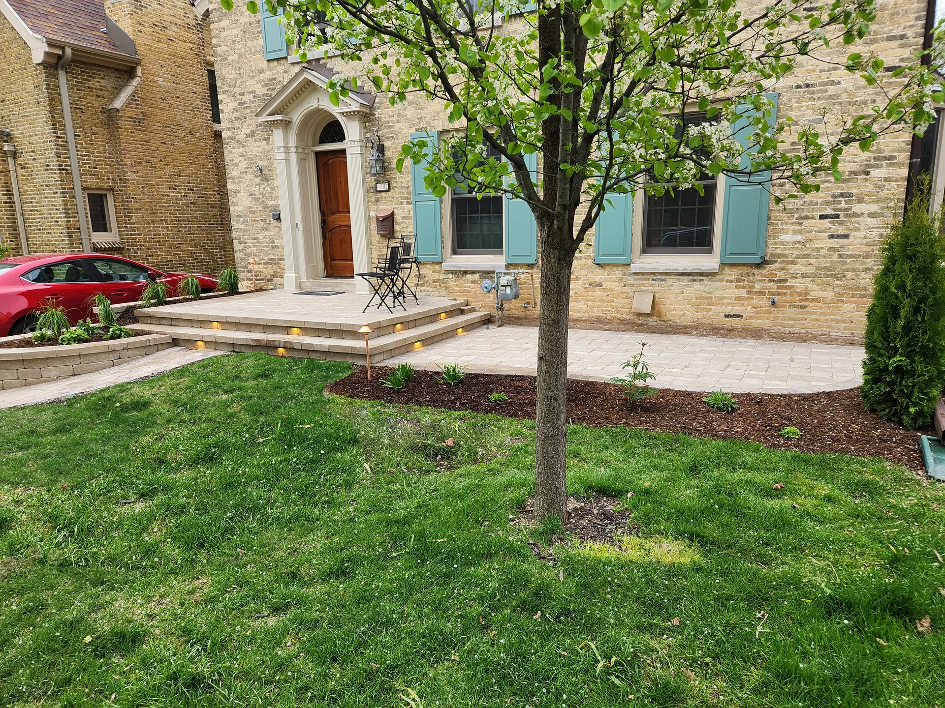 A tan stone house with blue shutters, a paved front patio, and a grassy lawn with a tree in the foreground.