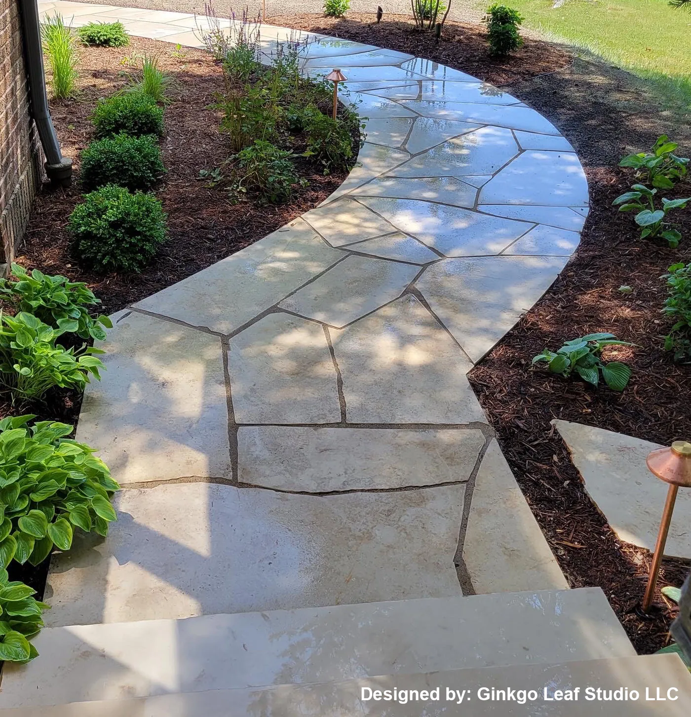 A curved flagstone pathway winds through a garden bed with lush green shrubs and dark mulch, viewed from above.