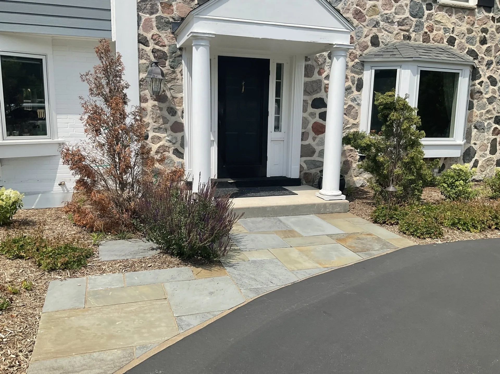A stone home exterior with a black front door under a white columned portico, flanked by shrubs and a stone walkway.