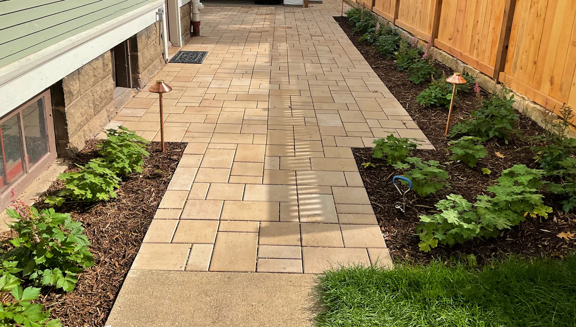 A stone paver walkway runs between a light green house wall and a wooden fence, flanked by garden beds with young plants.