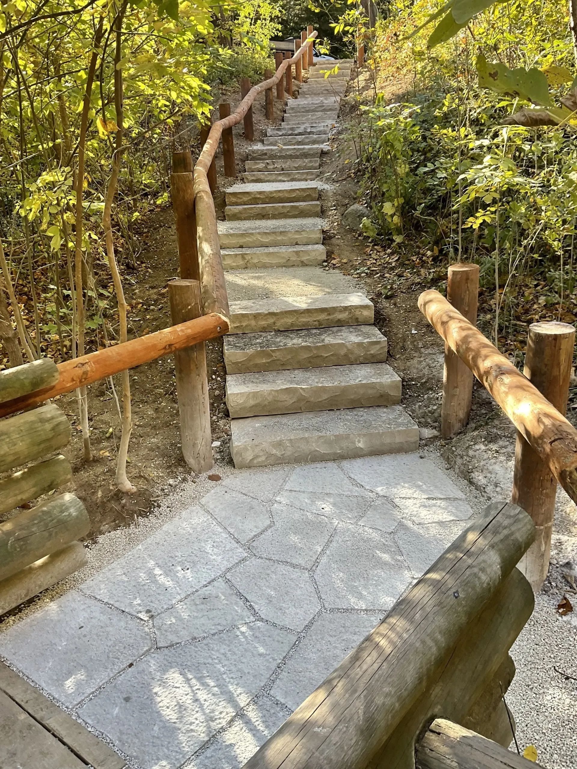 Stone steps lead up a wooded path, bordered by rustic wooden railings and a paved walkway in the foreground.