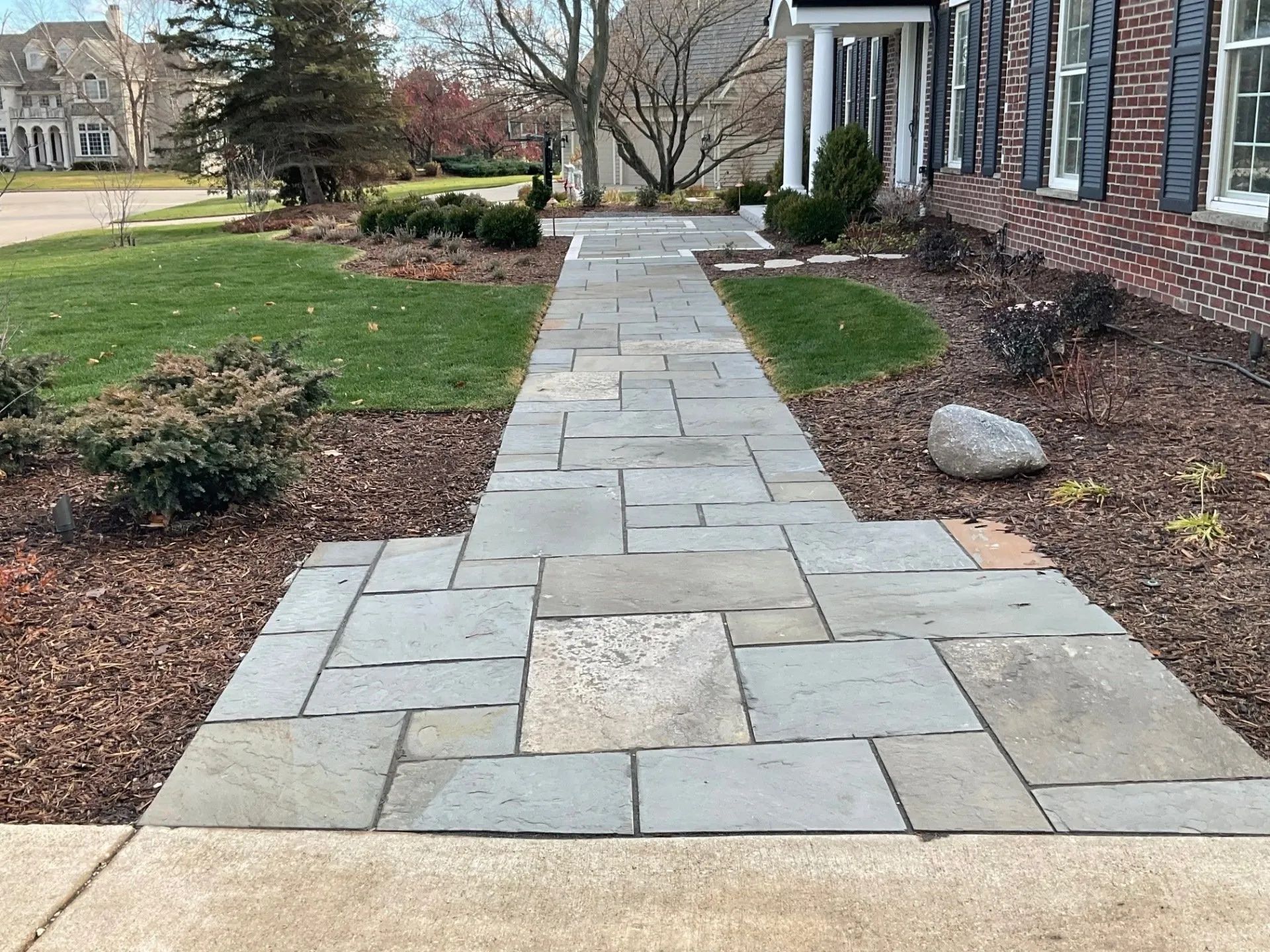 A stone walkway leads toward the entrance of a brick house, flanked by mulch beds, shrubs, and a green lawn.