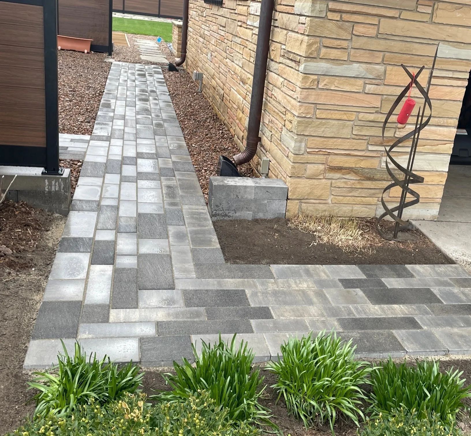 A gray paved walkway winds beside a tan stone house wall, a dark downspout, and a metal decorative garden sculpture.