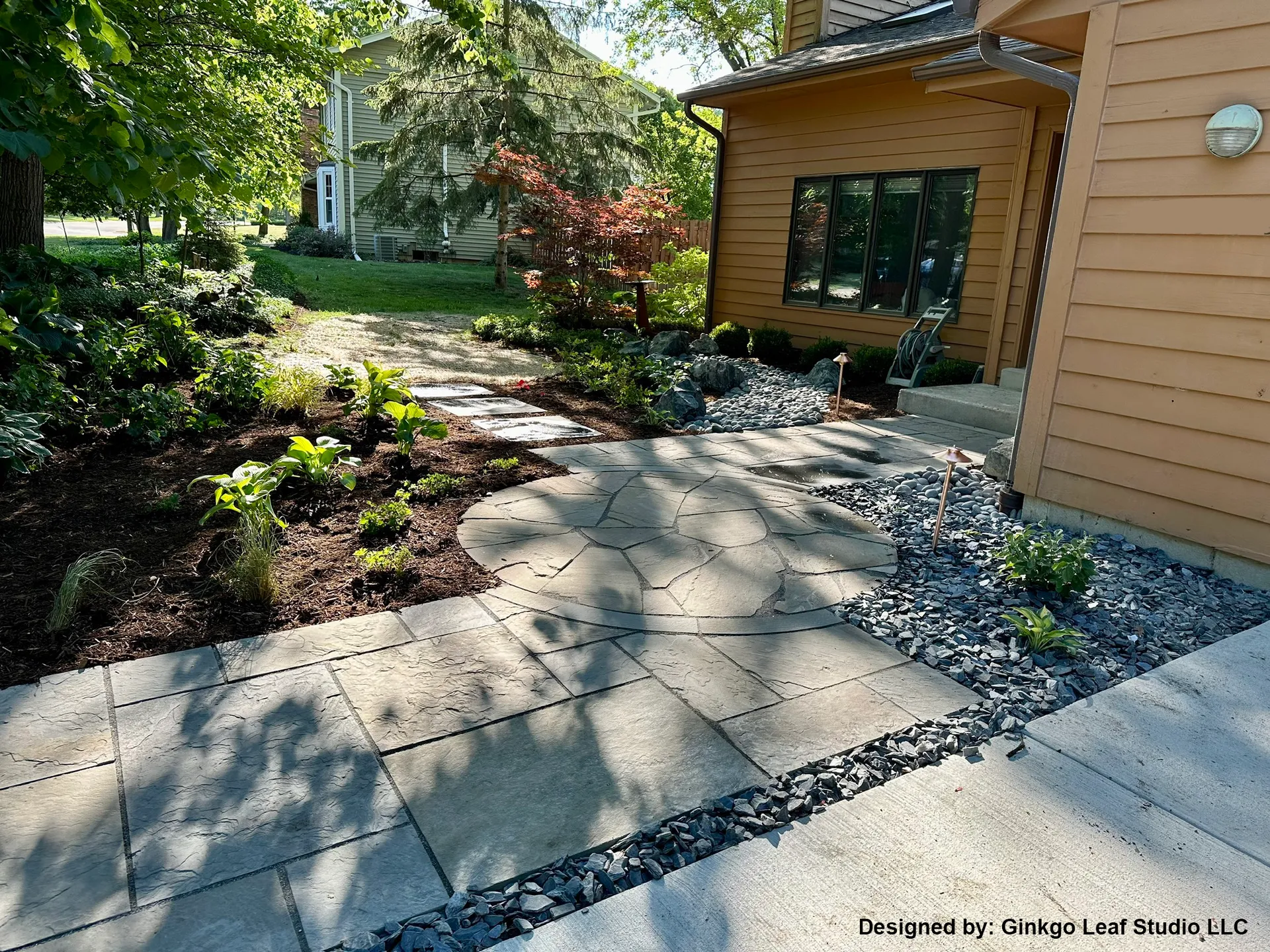 A landscaped front yard featuring a flagstone path, garden beds with mulch, and dark decorative stone near a tan home.